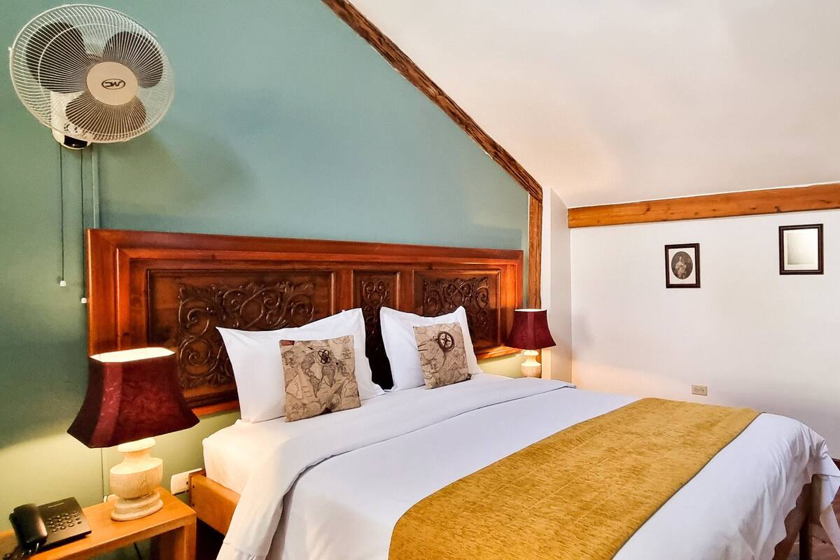 A bedroom with a wooden headboard, a bed with white bedding and a yellow throw, a bedside table with a lamp, a fan, and a TV on the wall.