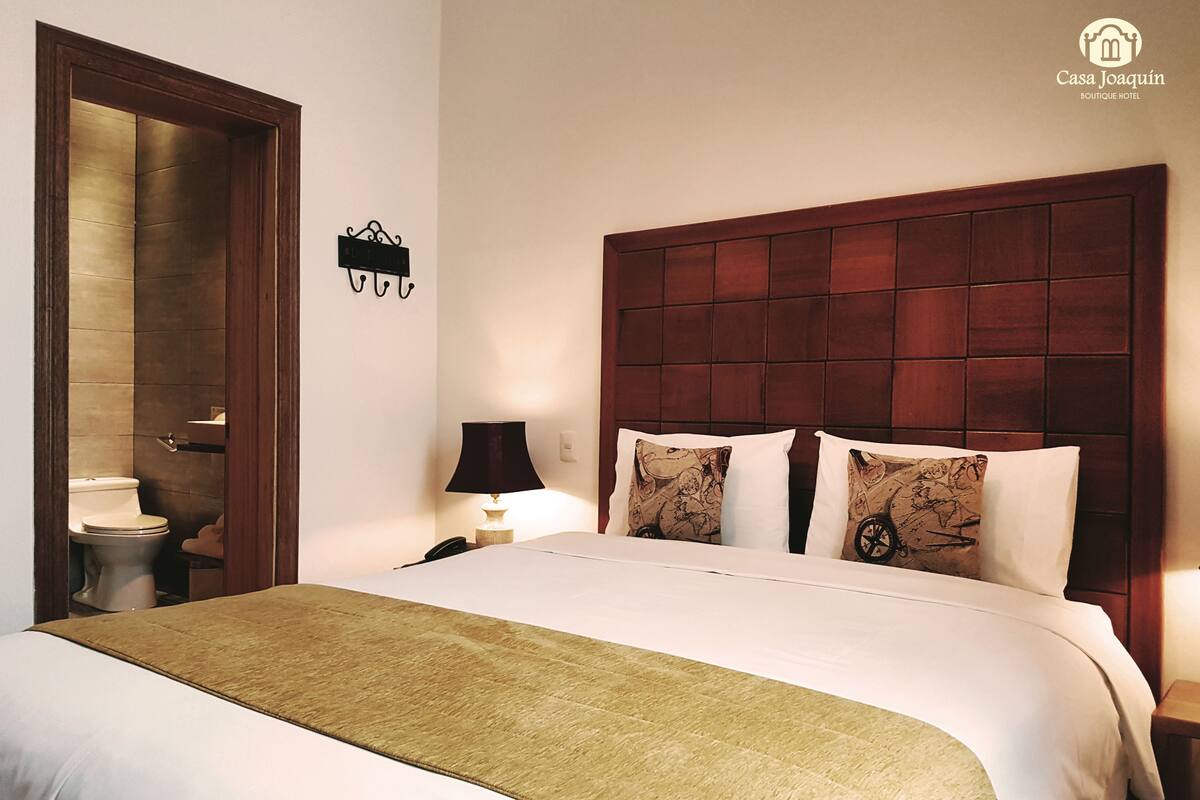A hotel room with a large bed, a wooden headboard, a nightstand with a lamp, and a bathroom visible through an open door.