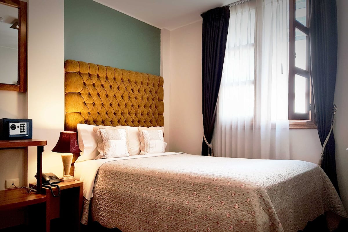 A hotel room with a tufted headboard, a bed with white linens, a bedside table with a lamp and a telephone, and a window with curtains.