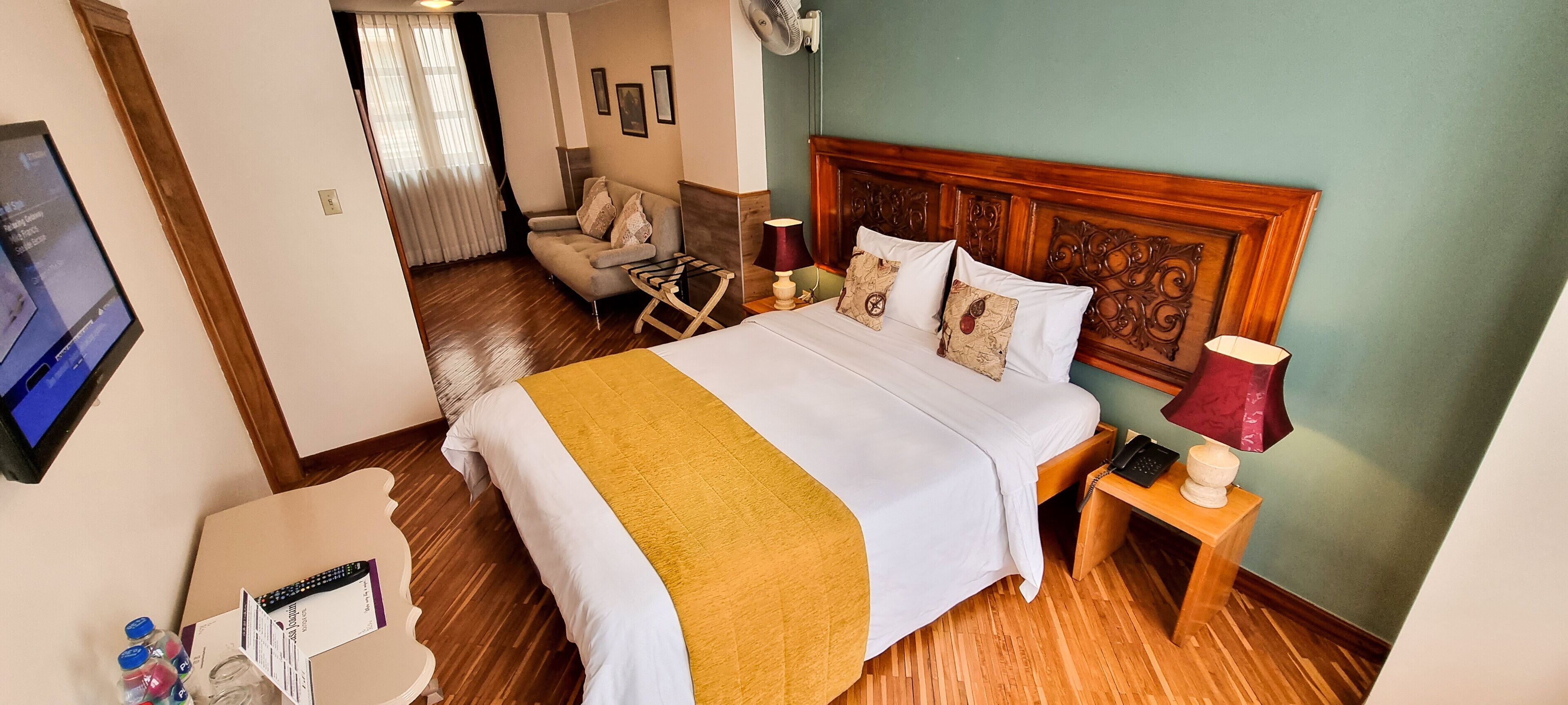 A hotel room with a bed, a wooden headboard, a nightstand with a lamp, a chair, and a television on the wall.
