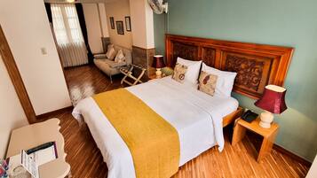 A hotel room with a bed, a wooden headboard, a nightstand with a lamp, a chair, and a television on the wall.