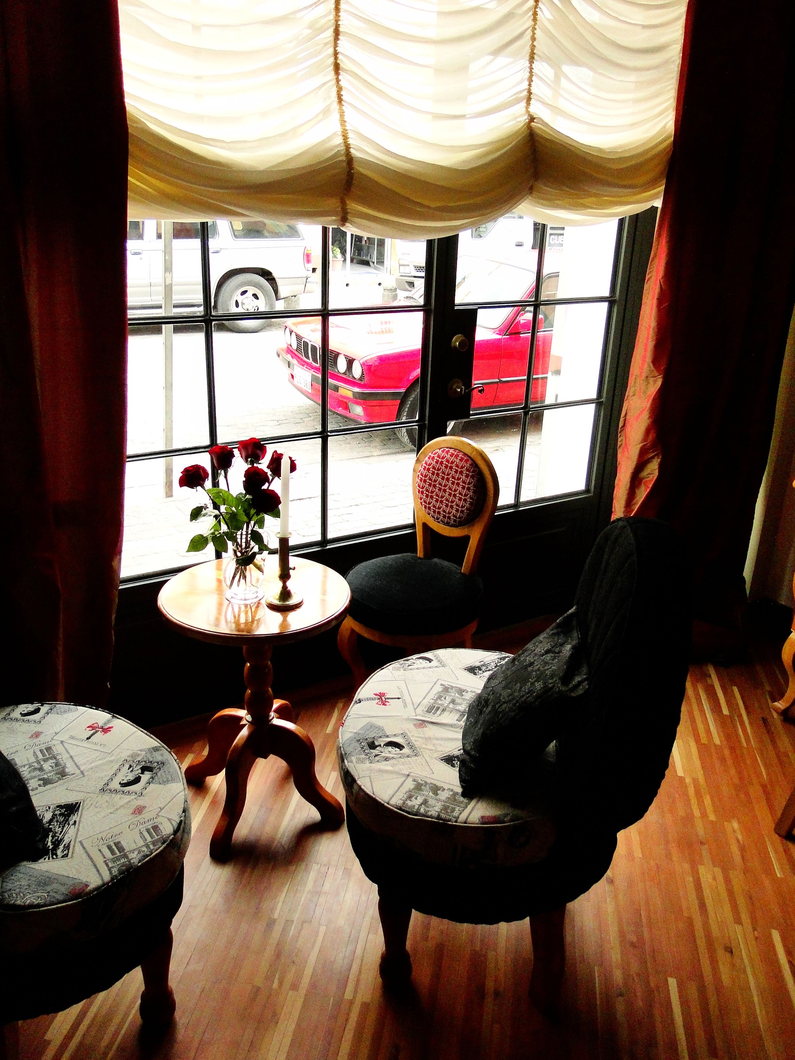 Una habitación con dos sillas, una mesita con un florero y vistas a un coche rojo a través de una ventana con cortinas.