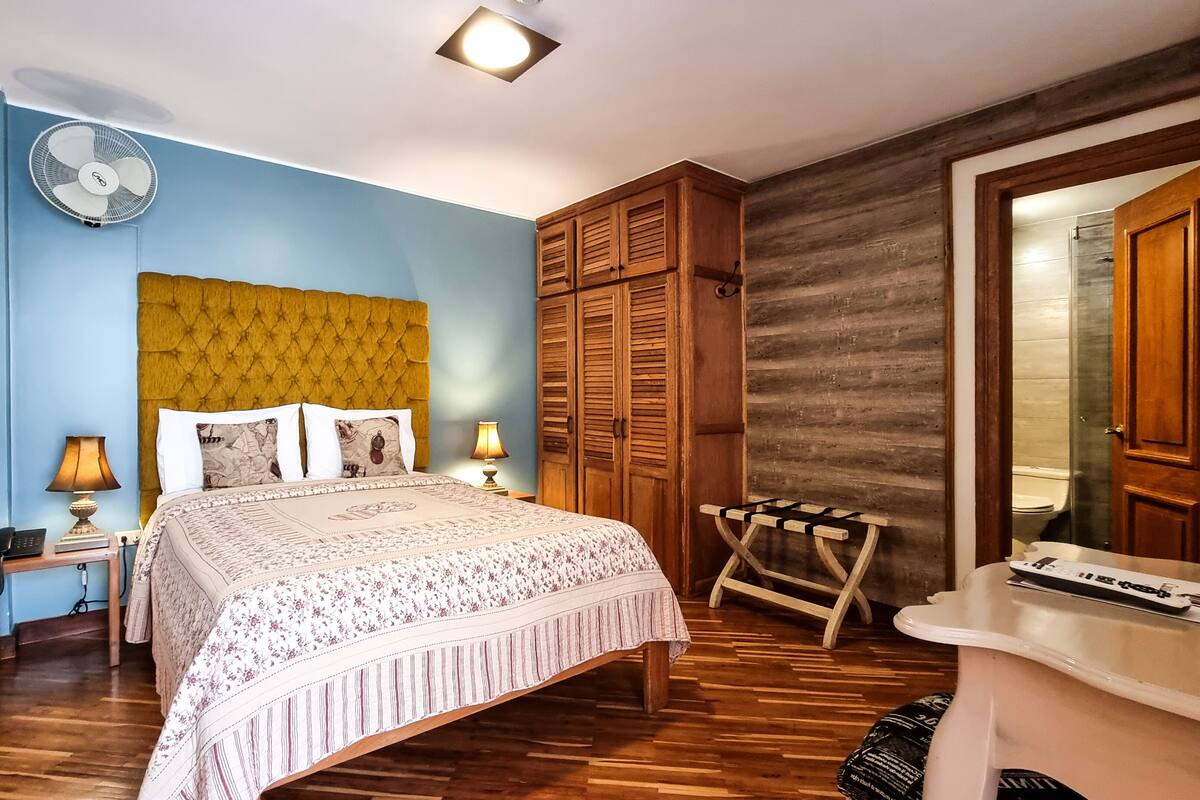 A bedroom with a bed, bedside lamps, a fan, a wooden wardrobe, and a small table.