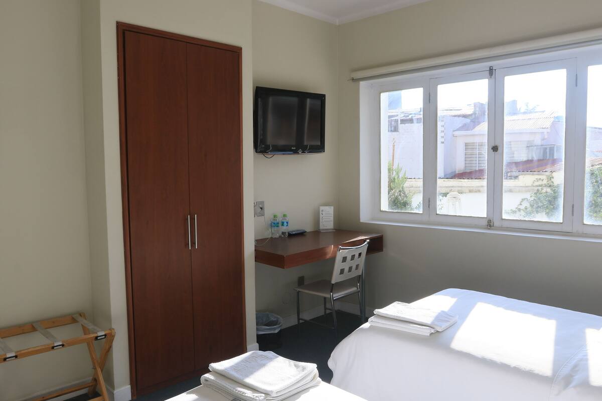 A hotel room with a wooden wardrobe, a desk with a chair, a bed with white linens, a television mounted on the wall, and a window with a view of buildings.