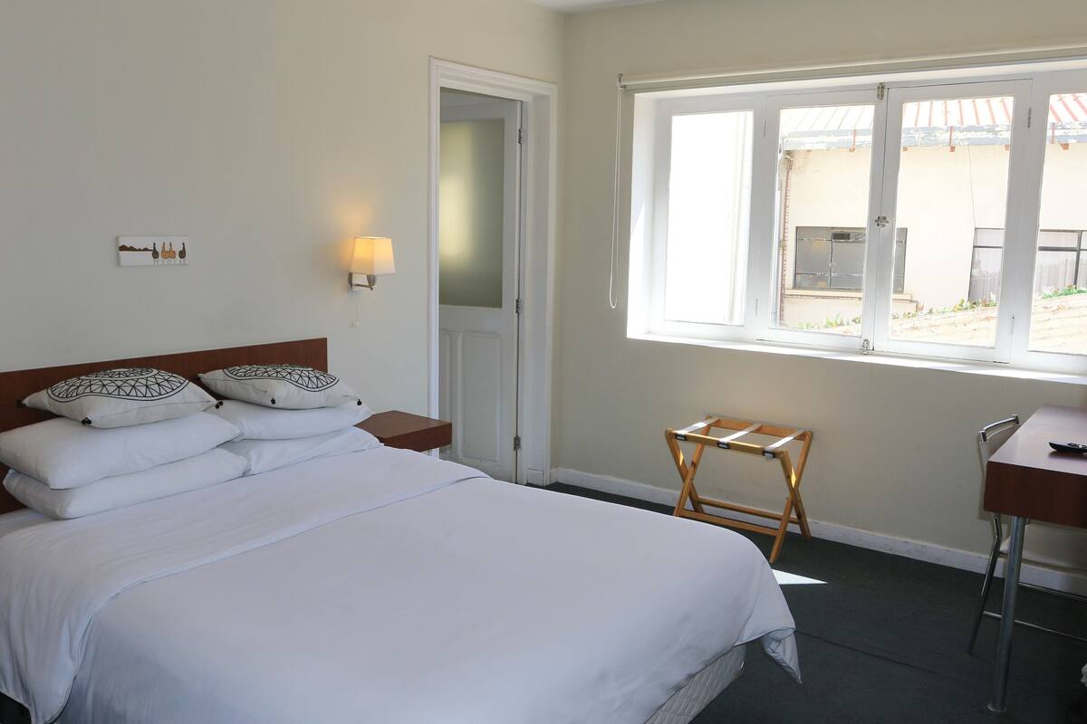 A neatly made bed with white linens and decorative pillows, a bedside table with a lamp, a wooden chair, and a window with a view of the outside.