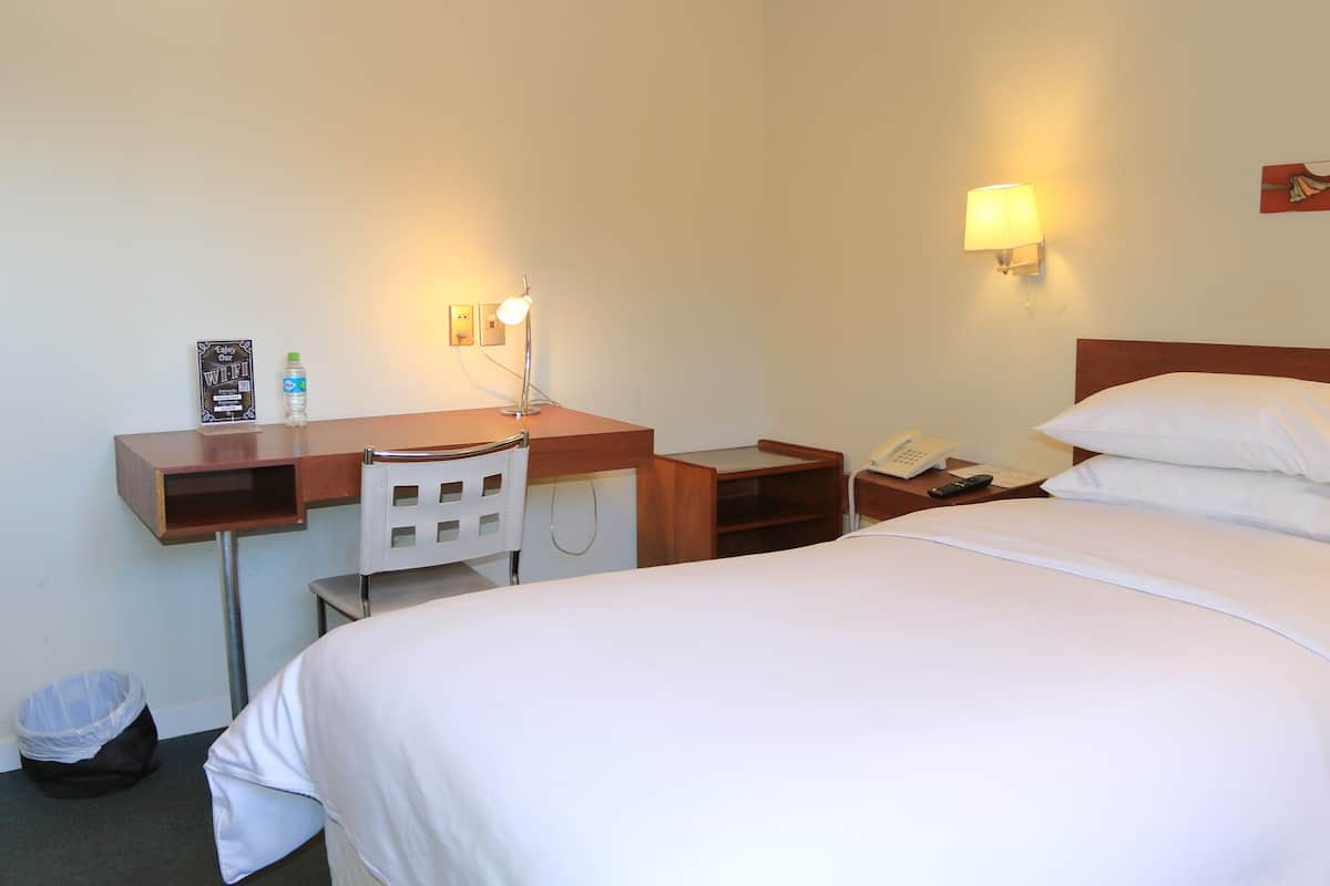 A hotel room with a bed, a desk, a chair, a lamp, a phone, and a trash bin.