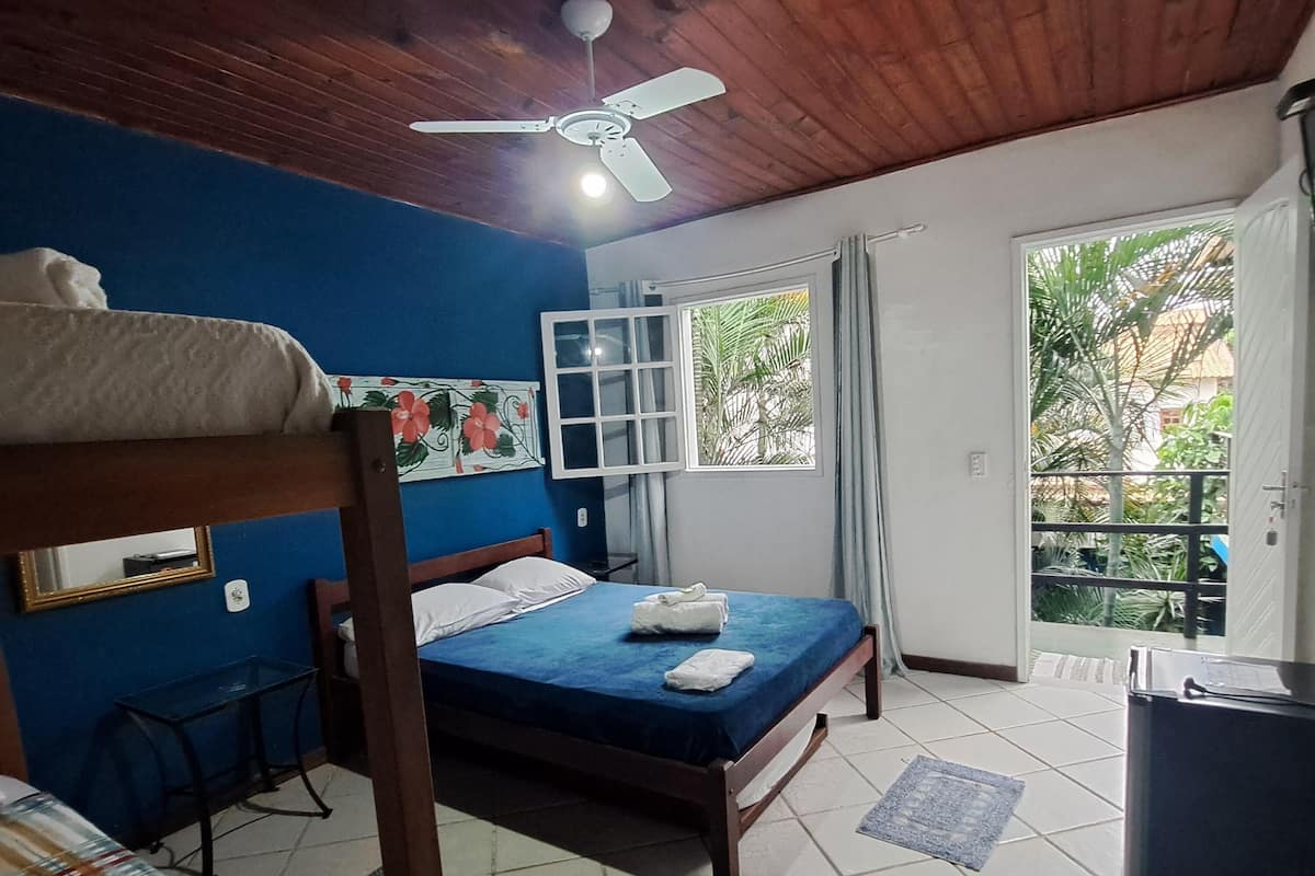 A room with a bed, a ceiling fan, a window with curtains, a desk, and a view of greenery outside.
