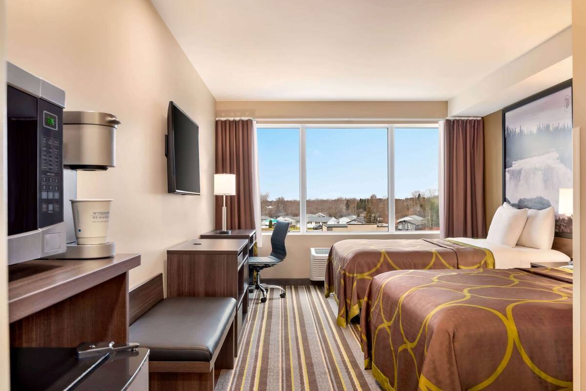 A hotel room with a desk, a chair, a bed with patterned bedding, a television, a microwave, and a window with a view of buildings.