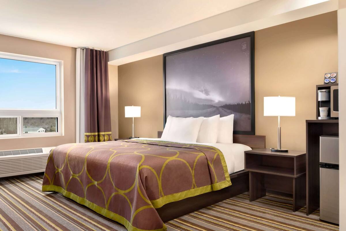 A hotel room with a large bed, two bedside tables, a nightstand, a refrigerator, a window with curtains, and a large framed picture on the wall.