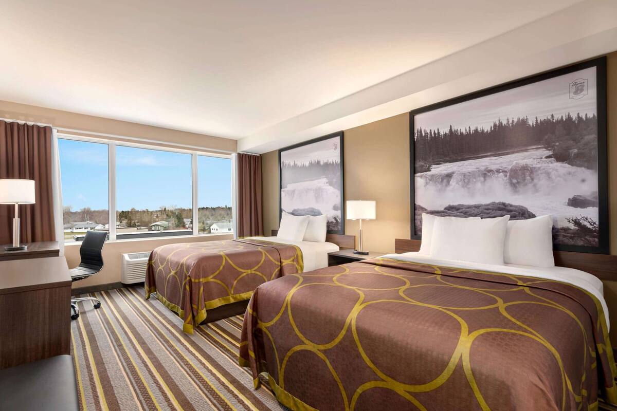 A hotel room with two beds, a desk, a chair, and large windows offering a view of a snowy landscape.