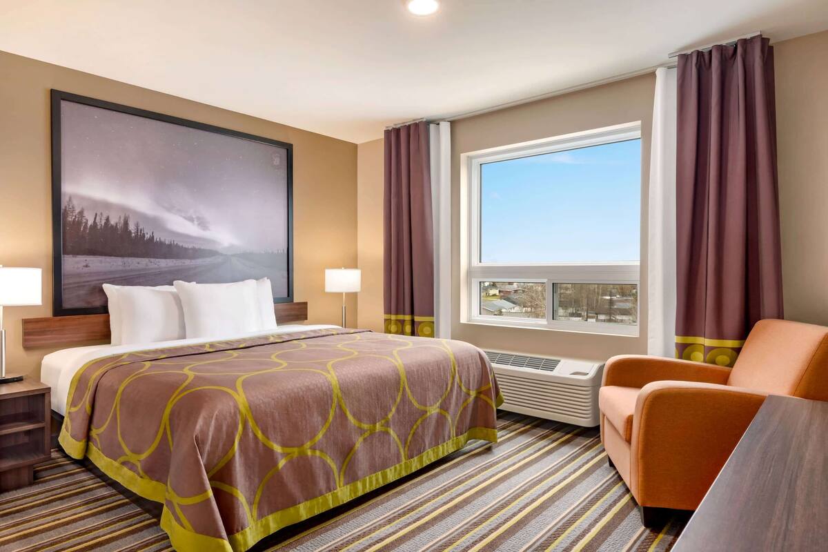 A hotel room with a large bed, a chair, a window with curtains, a wall-mounted air conditioner, and a large framed picture.