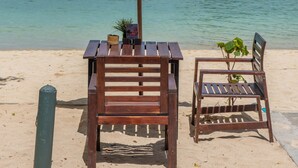 A beachside seating area with a table and chair under a large umbrella, overlooking a calm sea with boats and cliffs in the background. - PP Charlie Beach Resort (Ko Phi Phi)