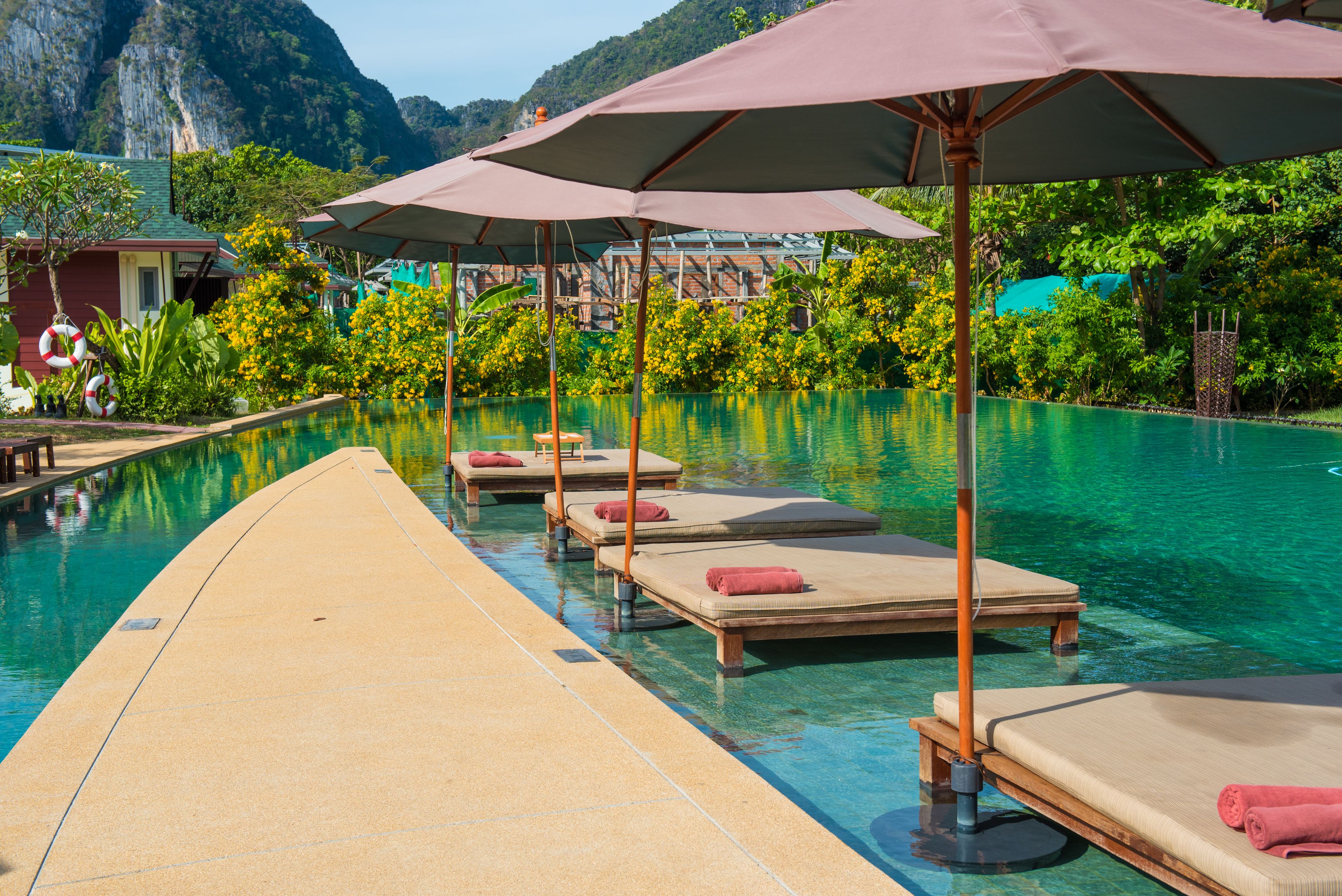 A clear swimming pool with lounge chairs and umbrellas, surrounded by lush greenery and mountains in the background.