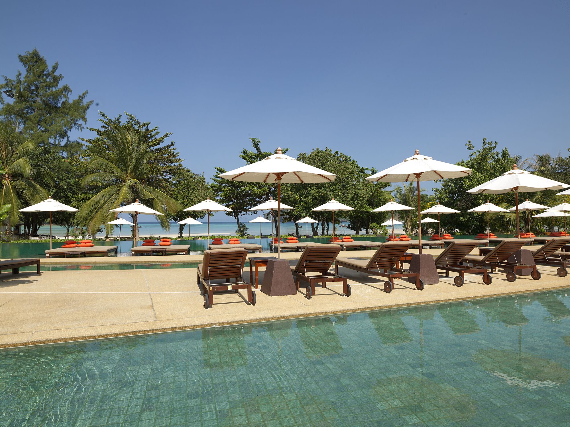 A poolside area with lounge chairs and umbrellas, surrounded by palm trees and a clear view of the ocean.
