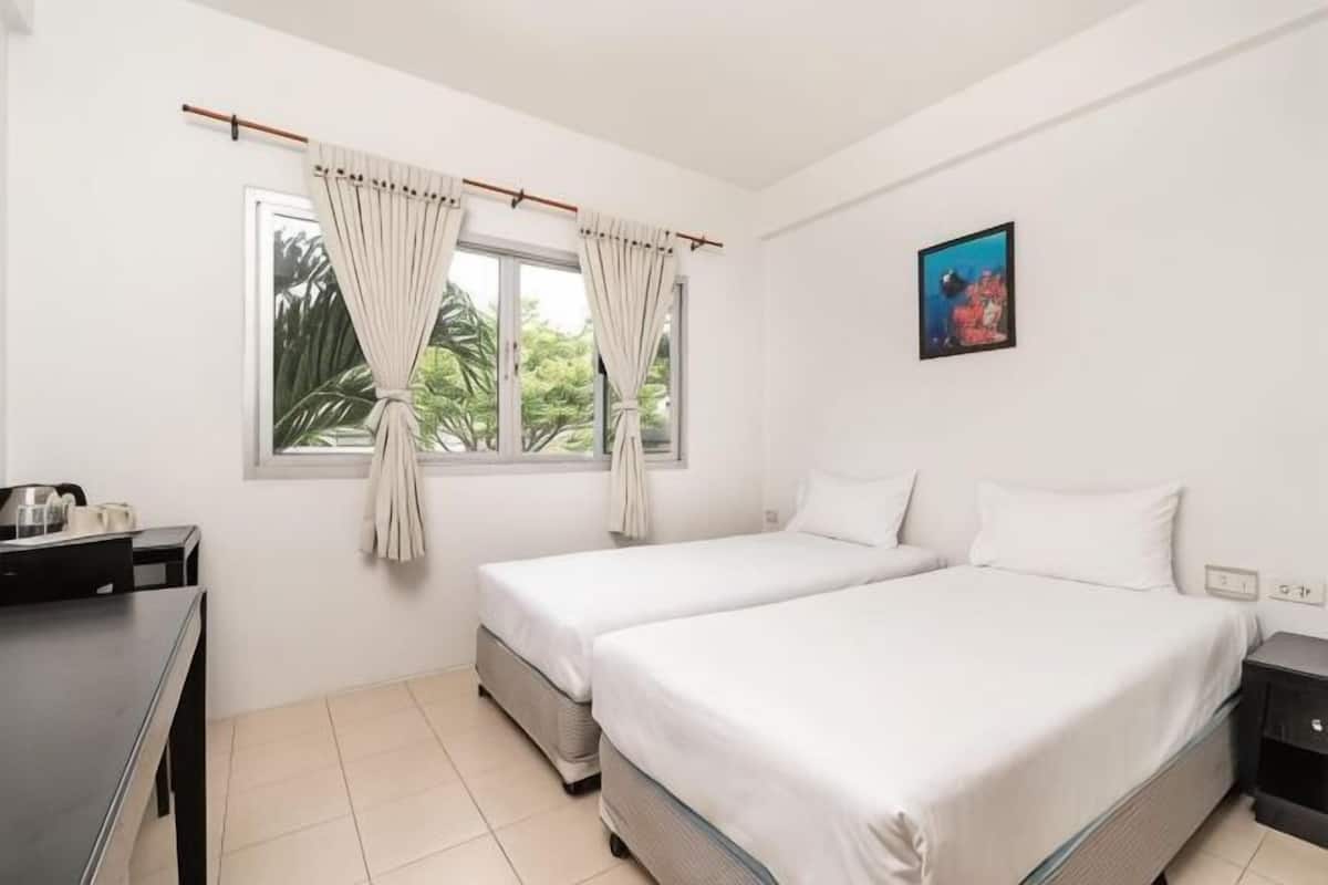 A room with two beds, a window with curtains, a desk with a chair, and a small table with a lamp.