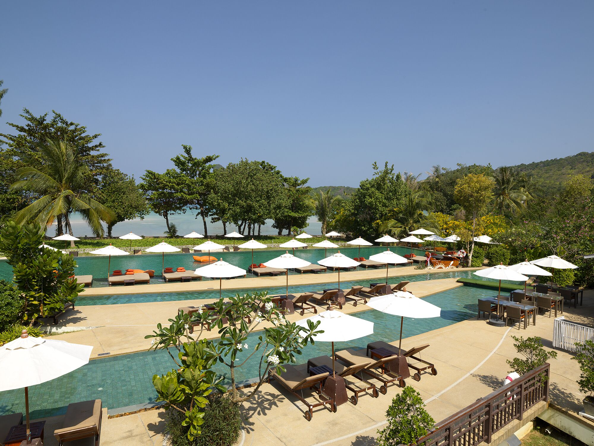 A pool area with multiple lounge chairs and umbrellas, surrounded by lush greenery and a clear view of the sea.