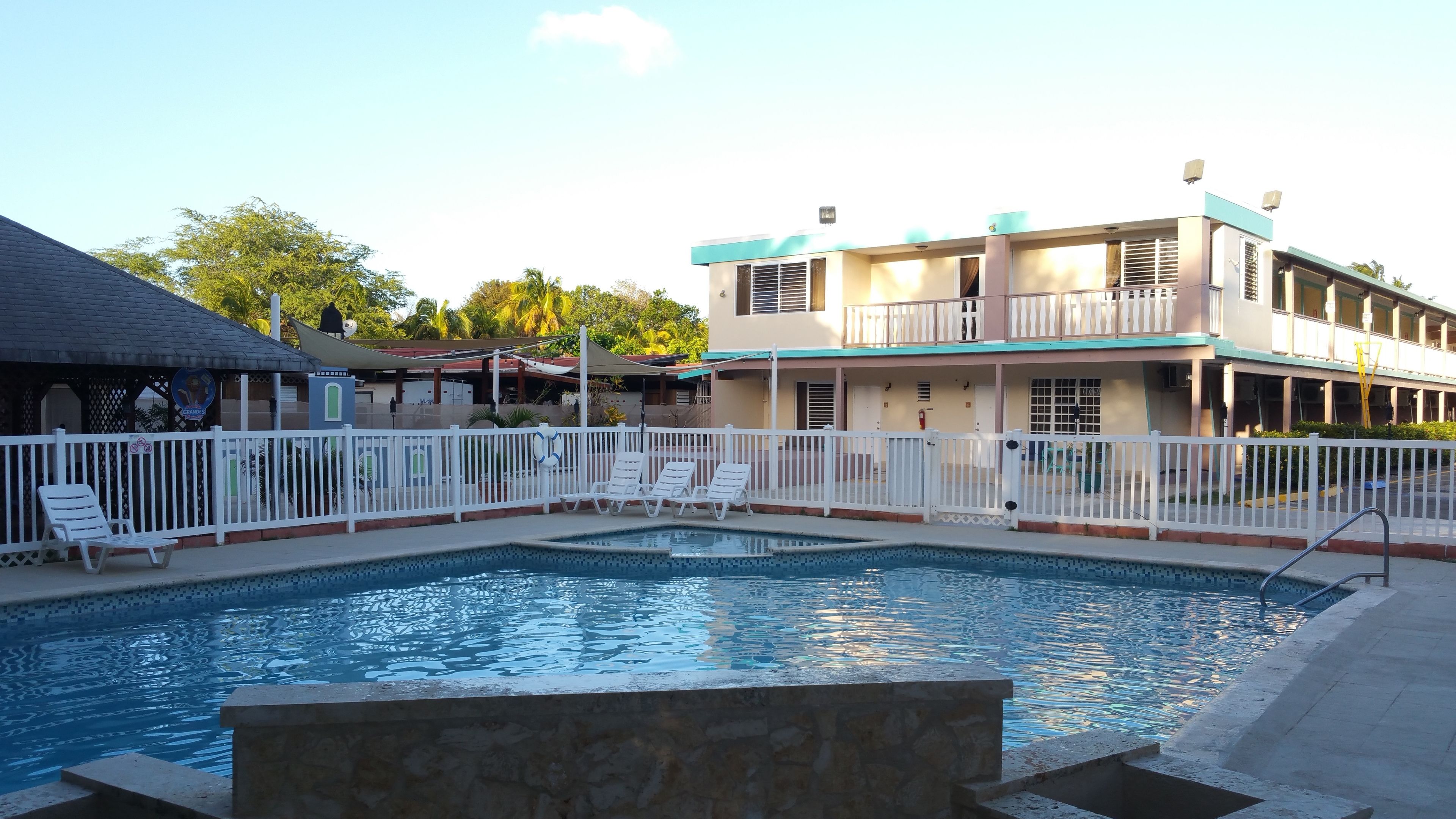 A swimming pool with white lounge chairs, a white fence, and a building with balconies in the background.
