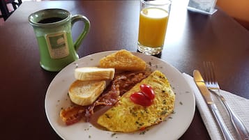Prato de pequeno-almoço com tosta, bacon, ovo e tomate, numa mesa com uma chávena verde e um copo de sumo de laranja.