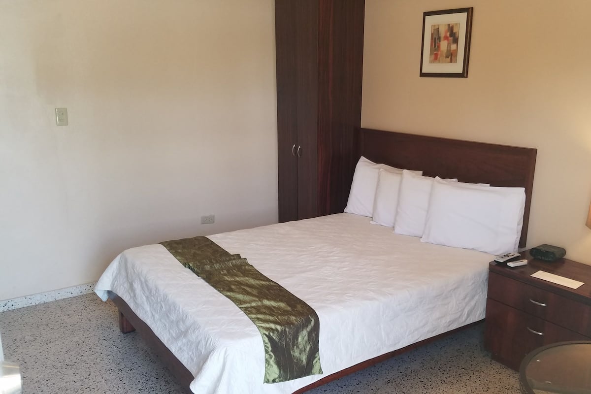 A neatly made bed with white linens and a green throw, a bedside table with a lamp, and a framed picture on the wall.