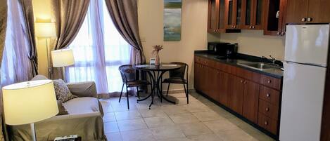 A compact living space with a sofa, a dining table with chairs, a kitchen area with wooden cabinets and a refrigerator, and a window with curtains.