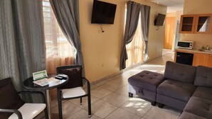 A living area with a sofa, a small table with a chair, a TV mounted on the wall, and a kitchen area with a microwave.