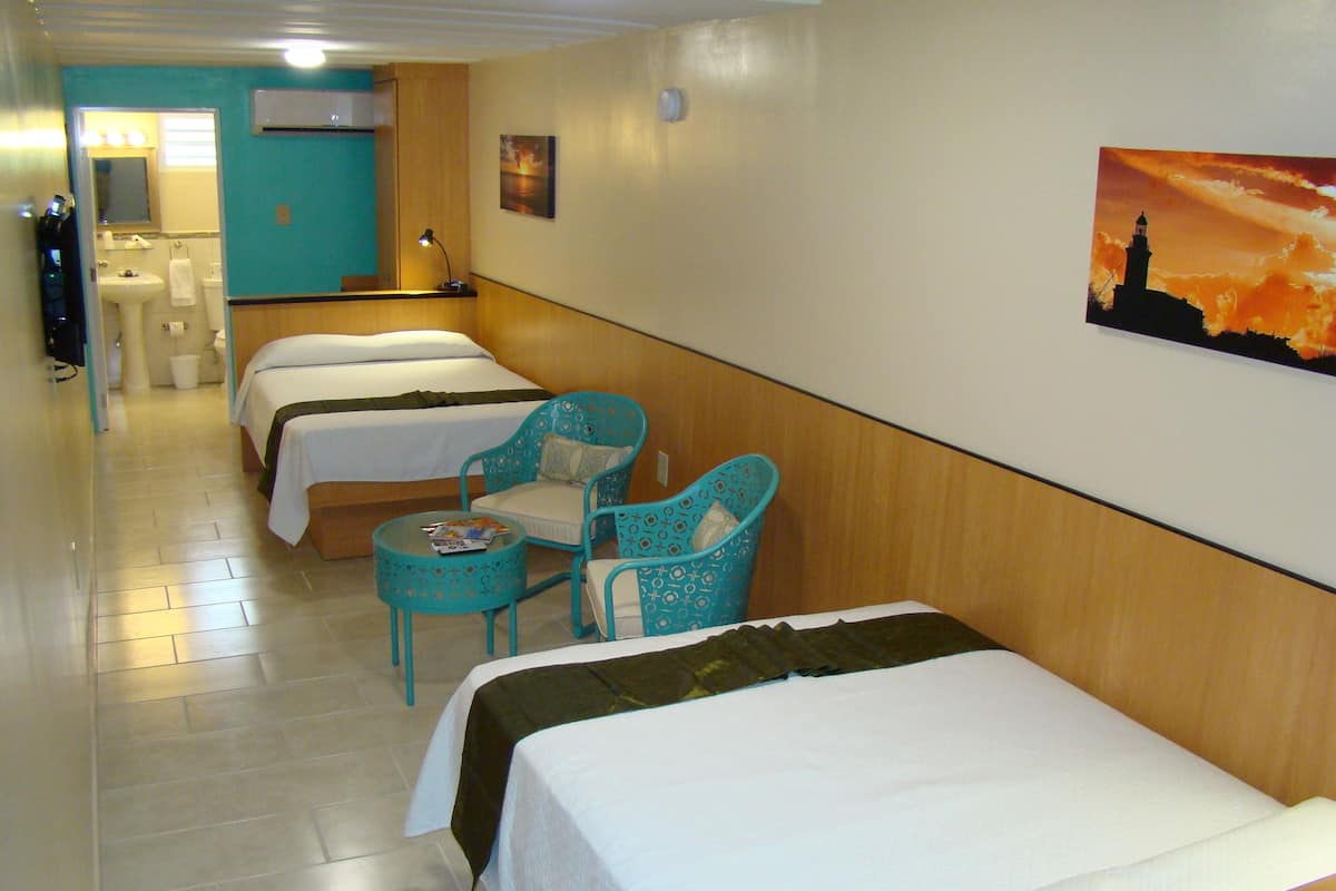 A hotel room with two single beds, a small table with a magazine, a chair, and a wall-mounted air conditioner.