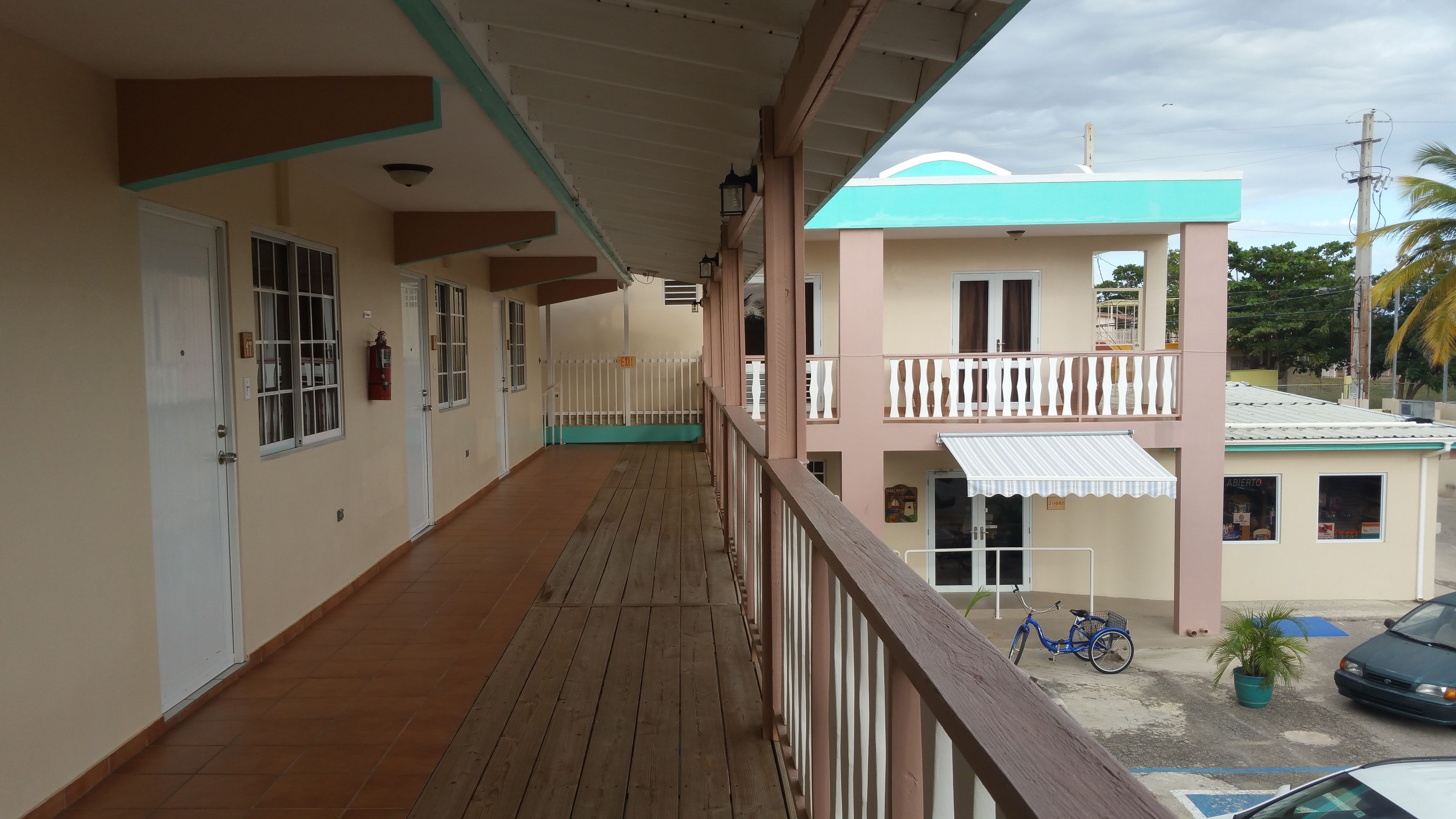 A covered outdoor corridor with a wooden floor, white railings, and a building with a balcony in the background.