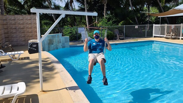 A person sitting on a swing over a swimming pool with a no diving sign, surrounded by palm trees and a fence.