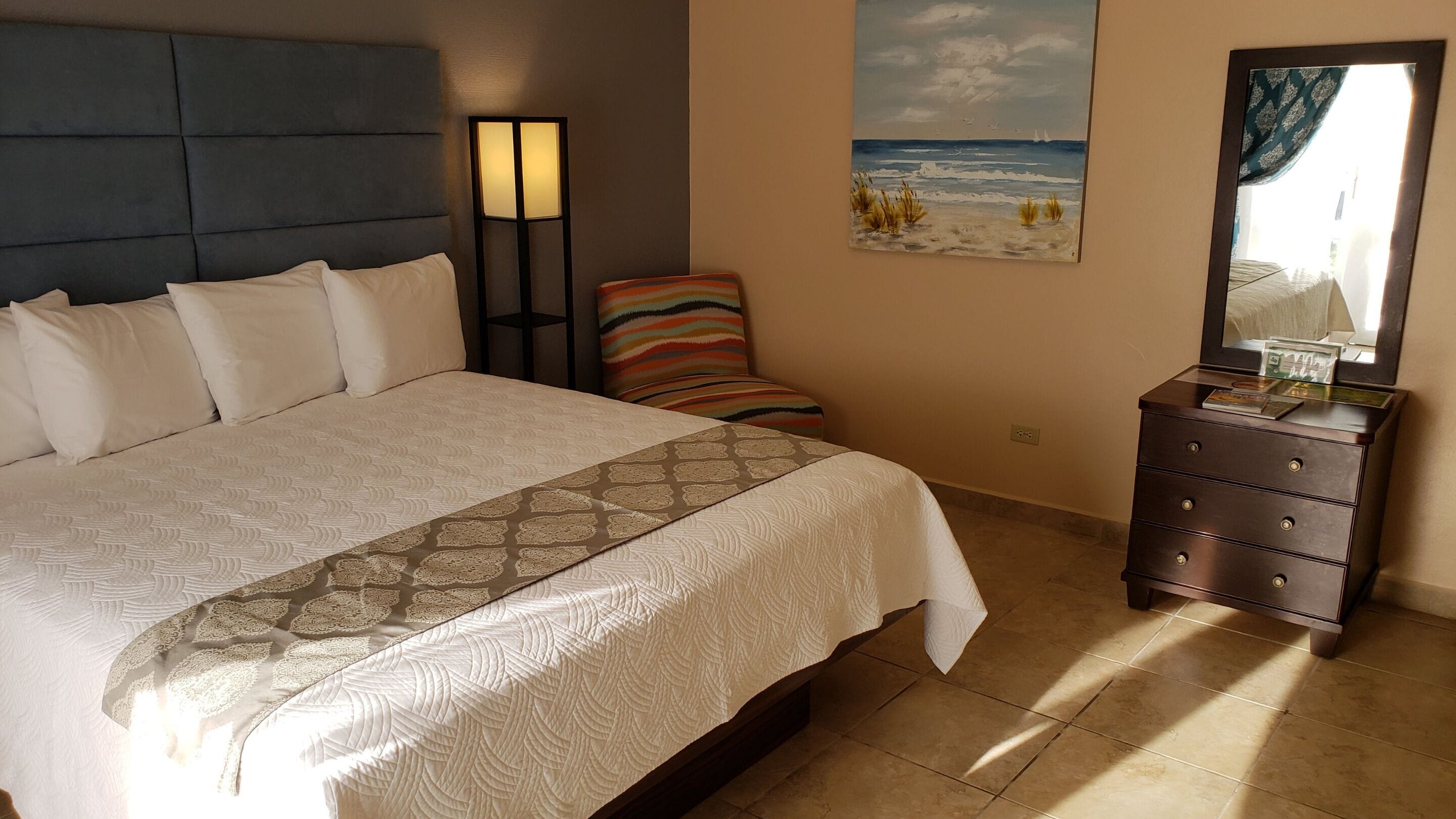 A neatly made bed with a patterned bedspread, a bedside lamp, a chair with a striped cushion, a dresser with a mirror, and a painting of a beach scene.