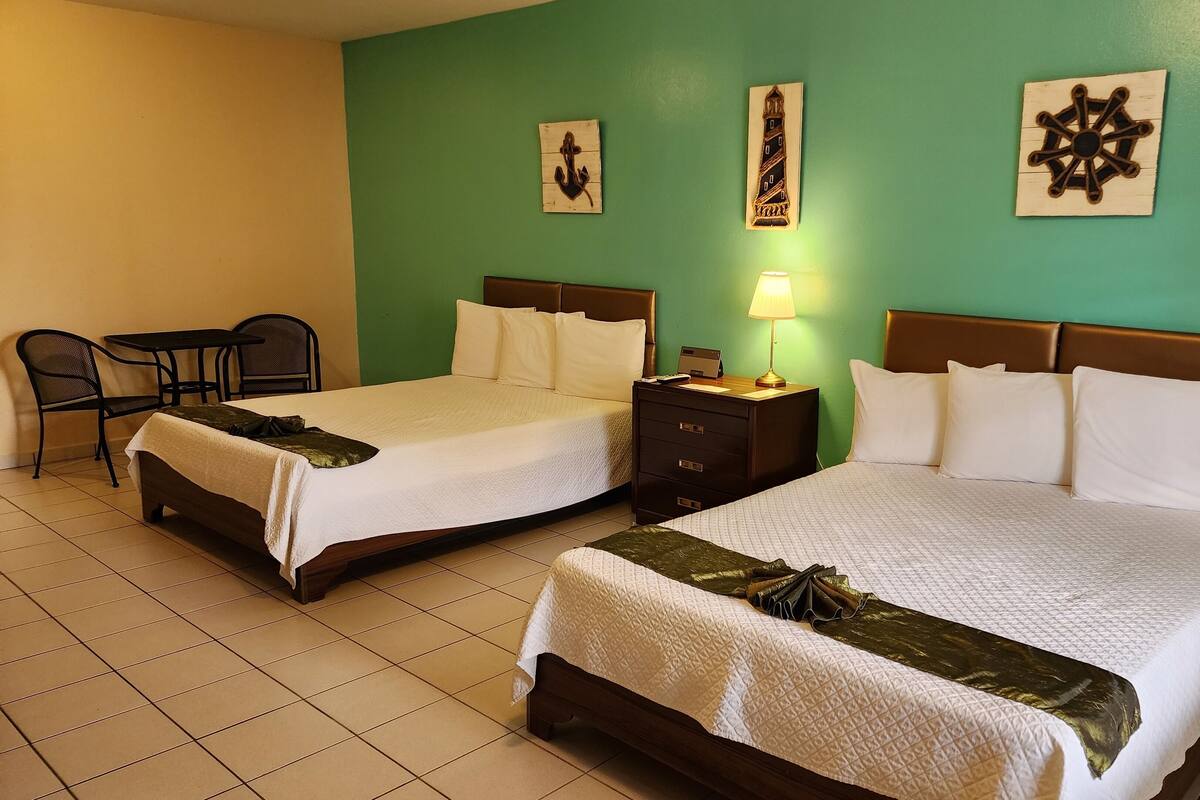 Two double beds with wooden headboards, a small table with chairs, a nightstand with a lamp, and wall art.