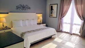 A hotel room with a large bed, two bedside tables with lamps, a nightstand, a painting on the wall, and a window with curtains.