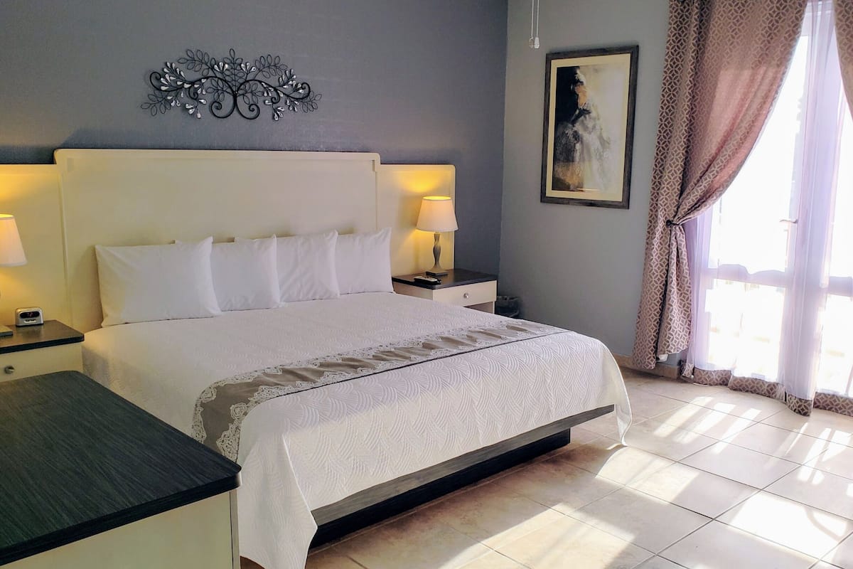 A hotel room with a large bed, two bedside tables with lamps, a nightstand, a painting on the wall, and a window with curtains.