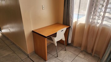 A small room with a wooden desk, a white chair, a window with sheer curtains, and tiled flooring.