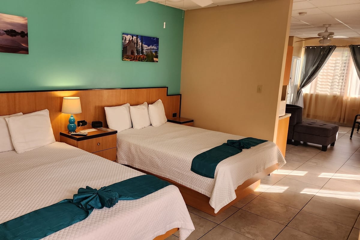 A hotel room with two beds, a nightstand with a lamp, a small table, and a chair.