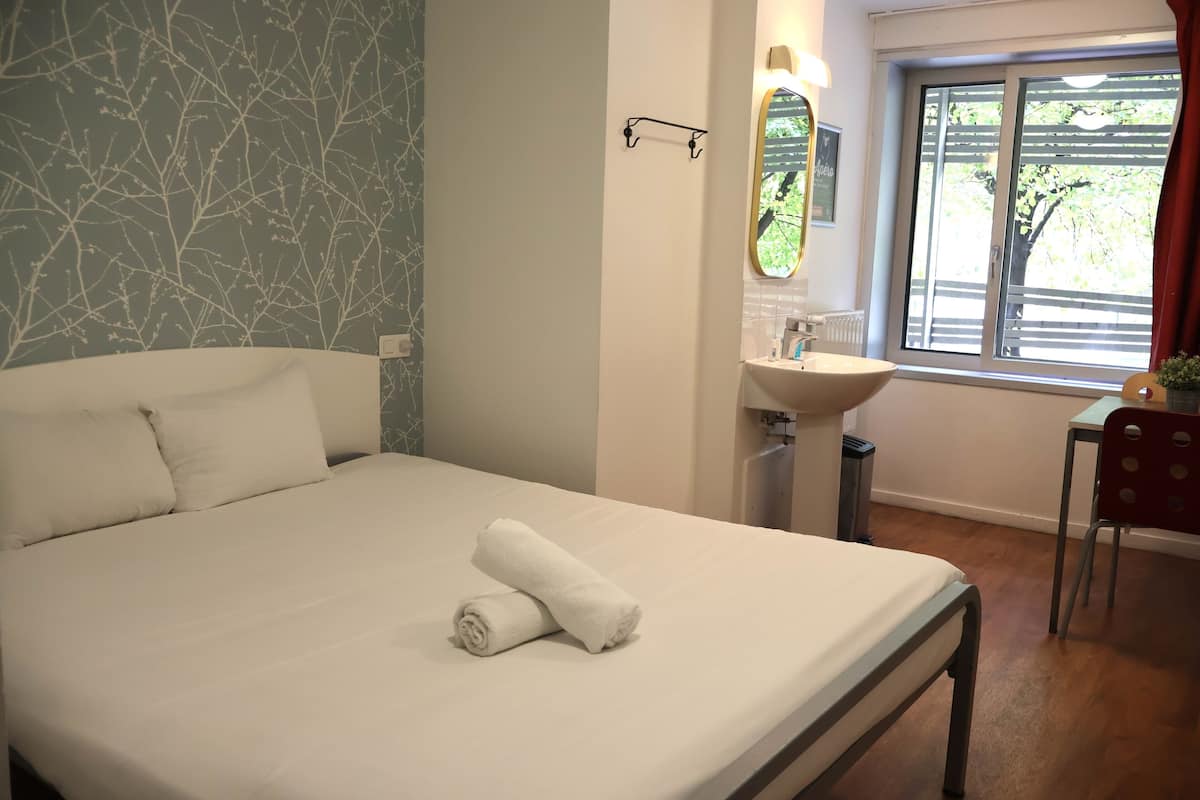 Una cama bien hecha con una toalla enrollada, una mesita de noche con una lámpara, una ventana con vistas a árboles, y una silla roja.