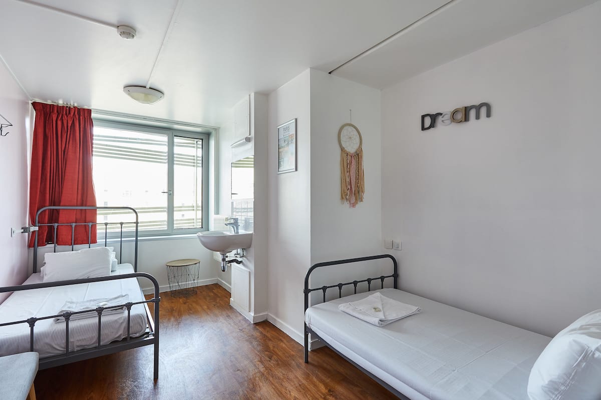 Un dormitorio con dos camas individuales, una ventana con cortinas rojas, un atrapasueños colgado en la pared y un lavabo blanco en la esquina.