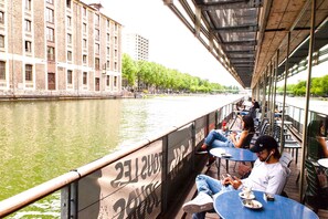 A riverside café with outdoor seating, a view of the river, and buildings in the background. - St Christopher's Inns The Canal Hostel (Paris)