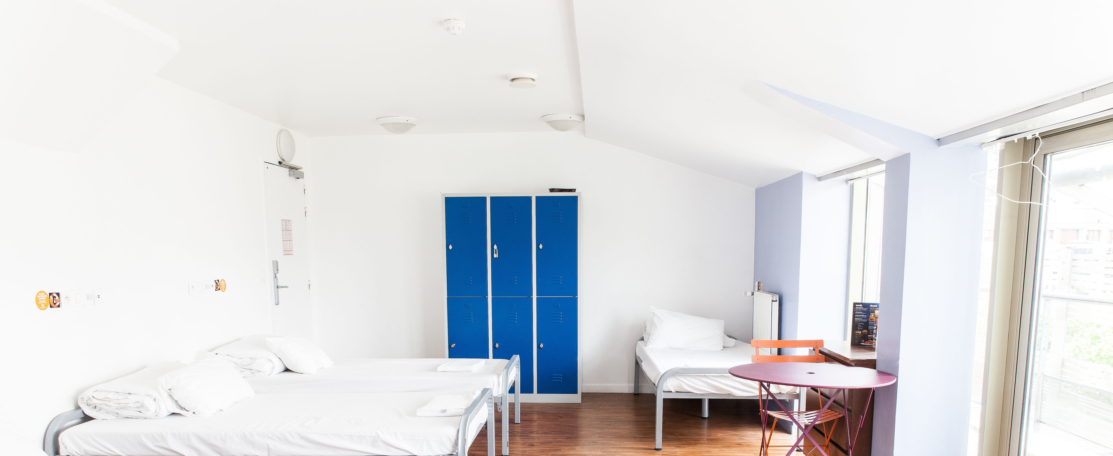 Habitació amb dos llits, un armari blau, una taula petita amb una cadira i una finestra.