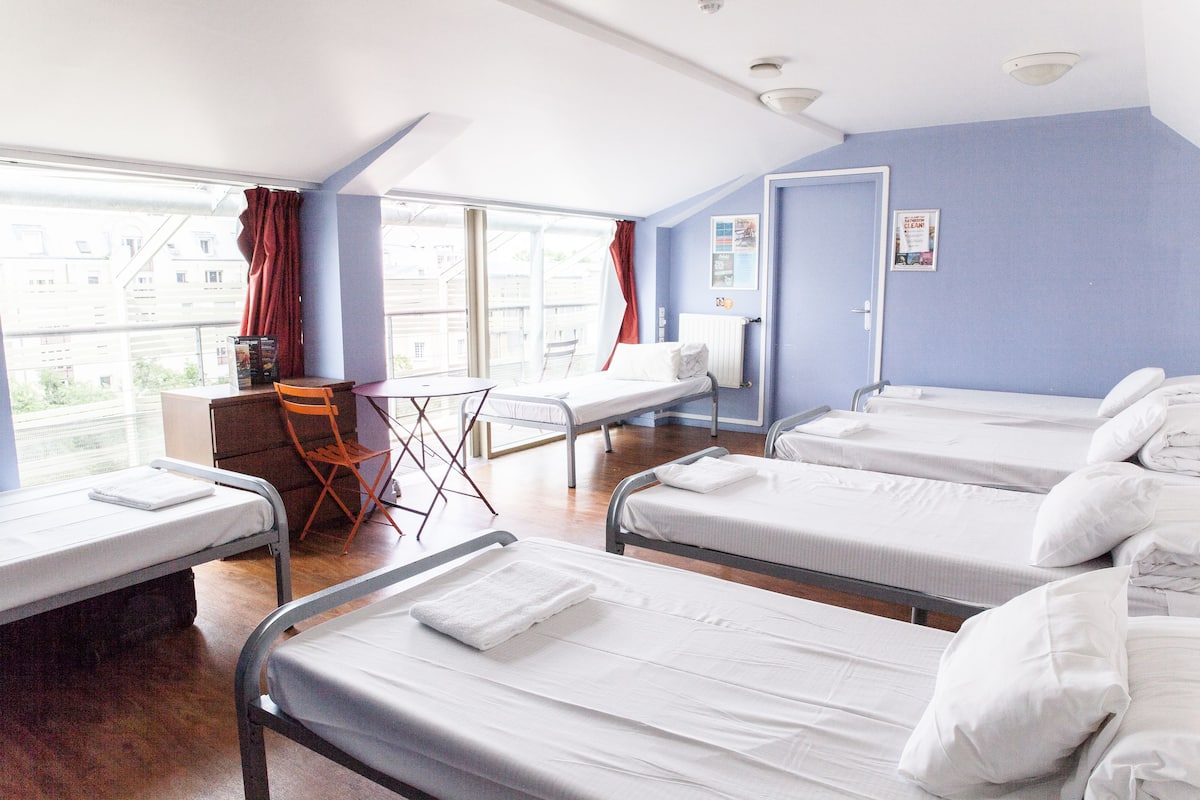 Habitación de hotel con cuatro camas individuales, un escritorio pequeño, una silla y una ventana con cortinas.