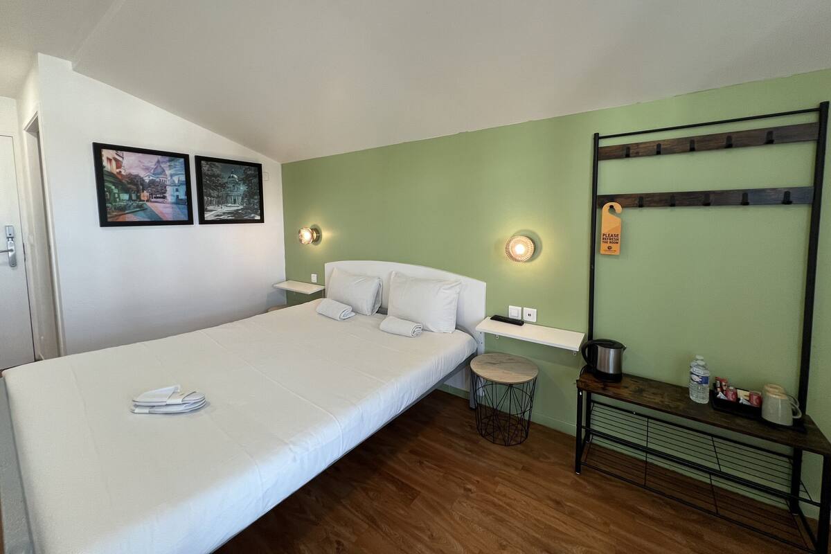 Un dormitorio con cama, dos lámparas fijadas a la pared, una mesita de noche con hervidor, una pequeña mesa redonda y un banco con ganchos.