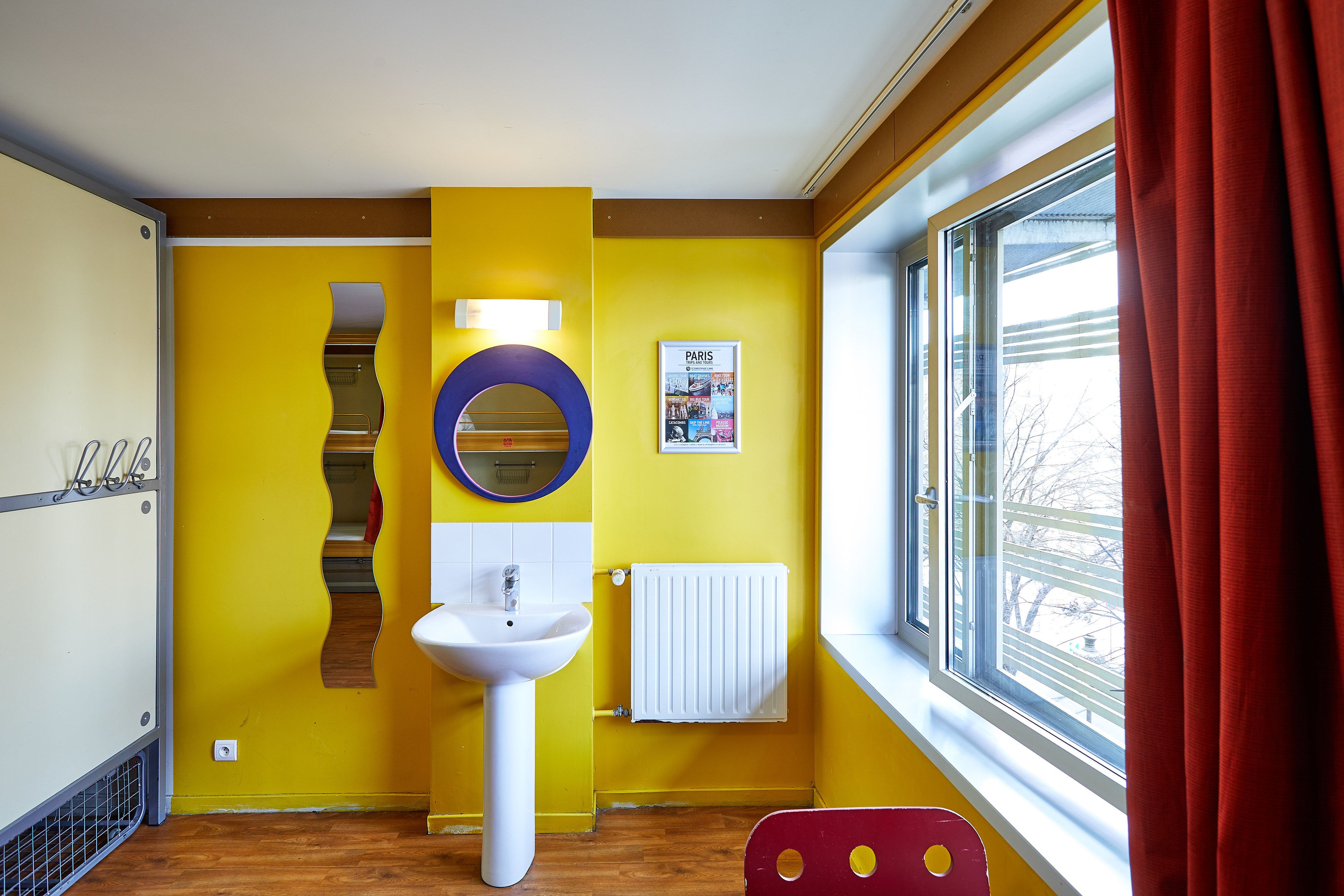 A bathroom with a yellow wall, a white sink, a mirror, a poster, and a window with a red curtain.
