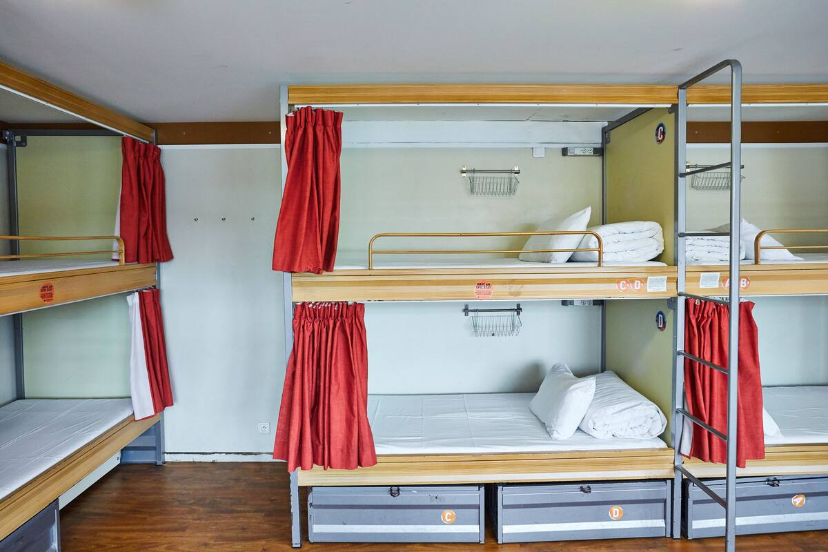 Habitación con literas, tres en total, cada una con una escalera metálica y una cortina roja. Hay cajones de almacenamiento debajo de las camas.
