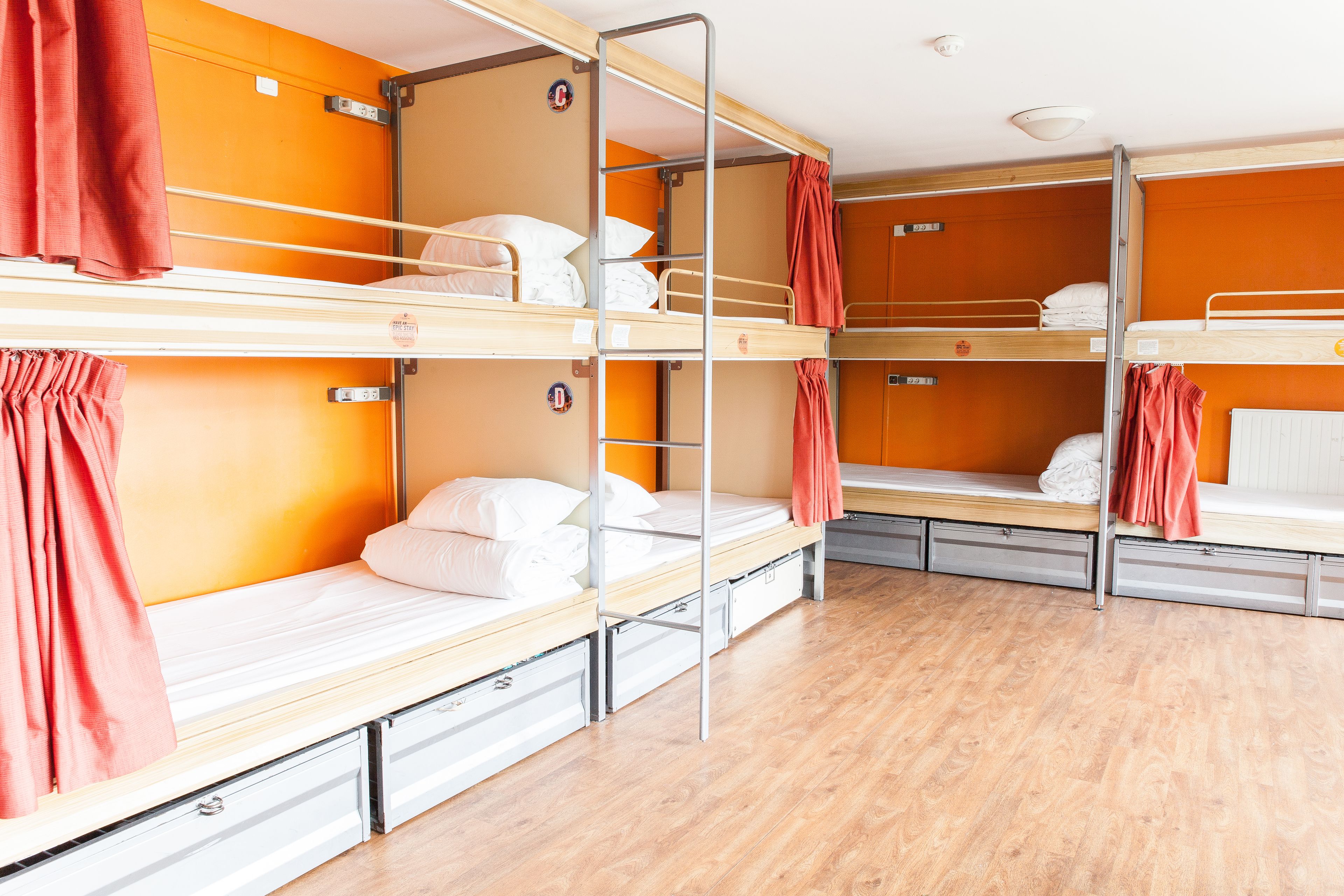 A dormitory with bunk beds, each with a mattress, pillows, and a red curtain. There are storage drawers under the beds.