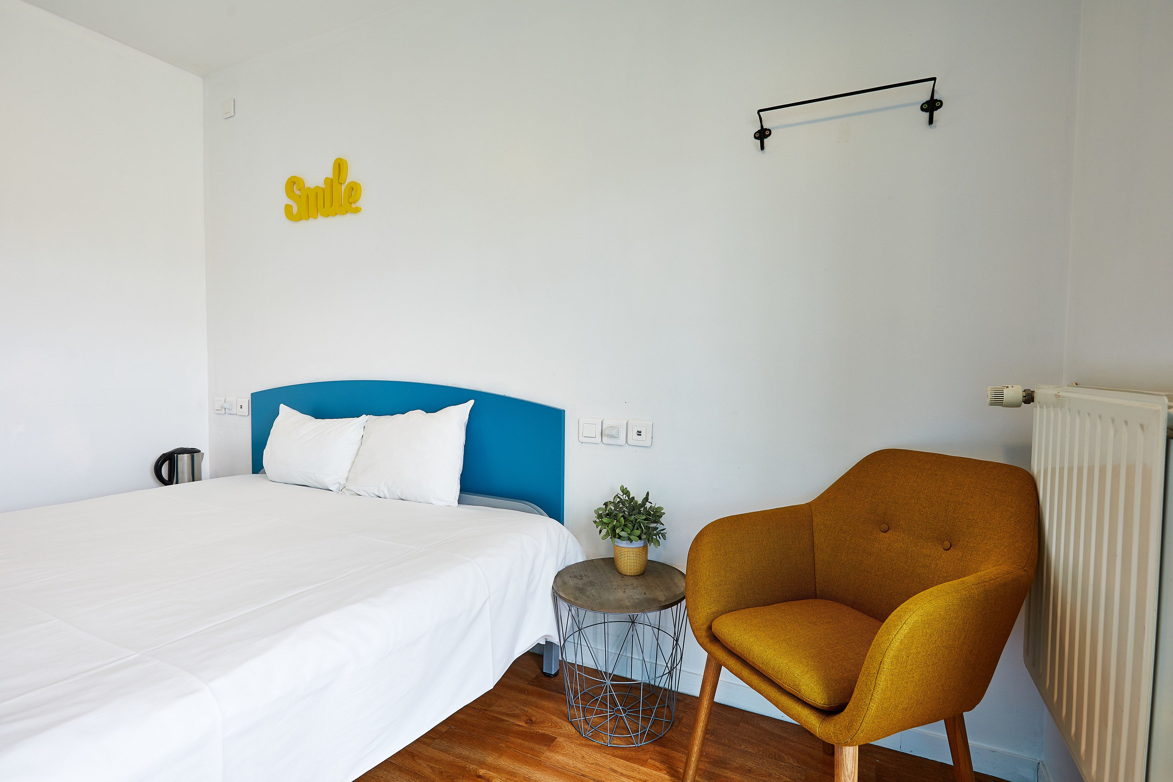 A hotel room with a bed, a blue headboard, a mustard-colored armchair, a small side table with a plant, and a wall-mounted light fixture.