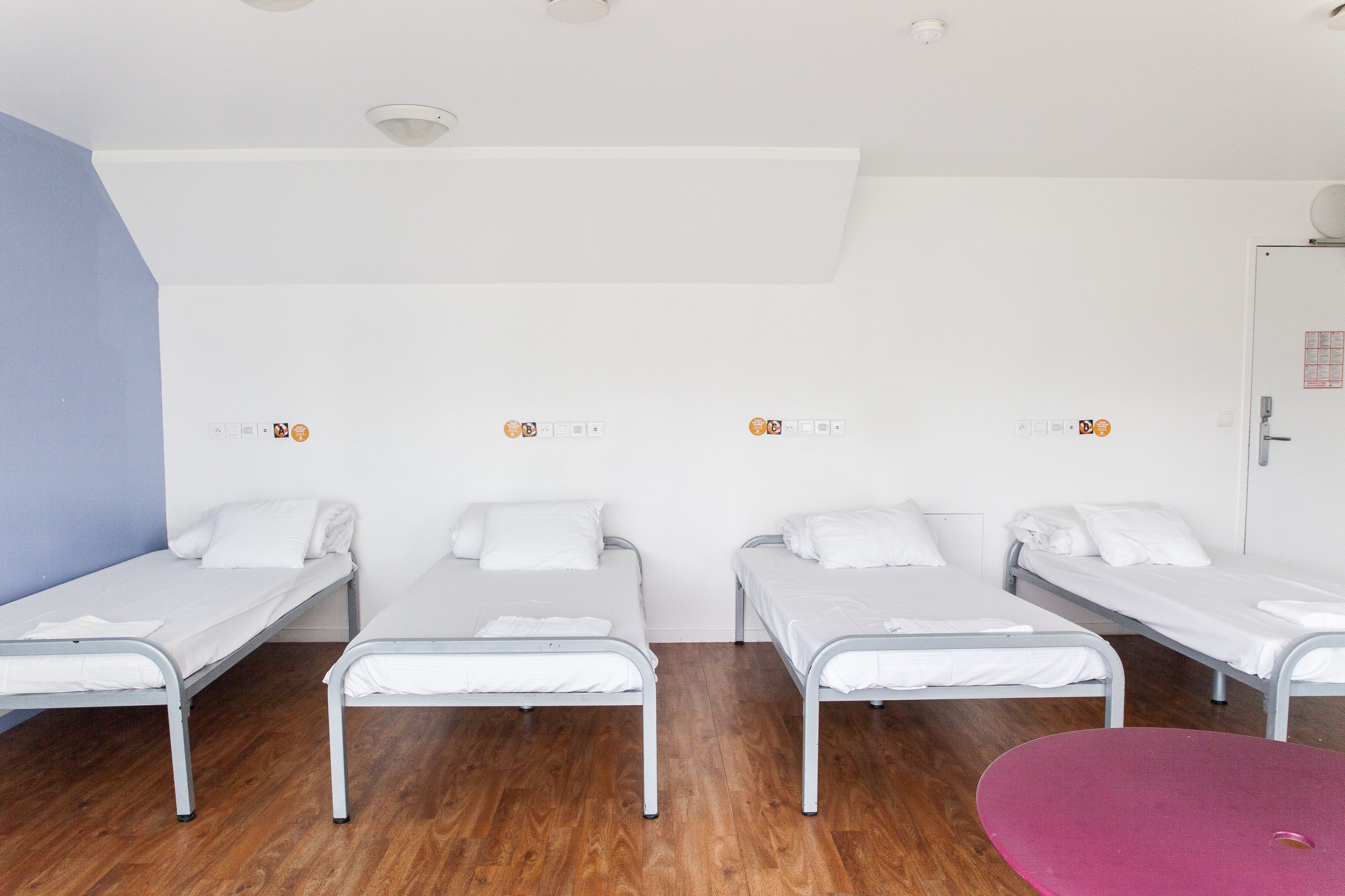 A room with four single beds aligned against a wall, a wooden floor, and a door on the right.