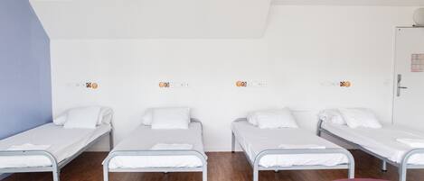 A room with four single beds aligned against a wall, a wooden floor, and a door on the right.