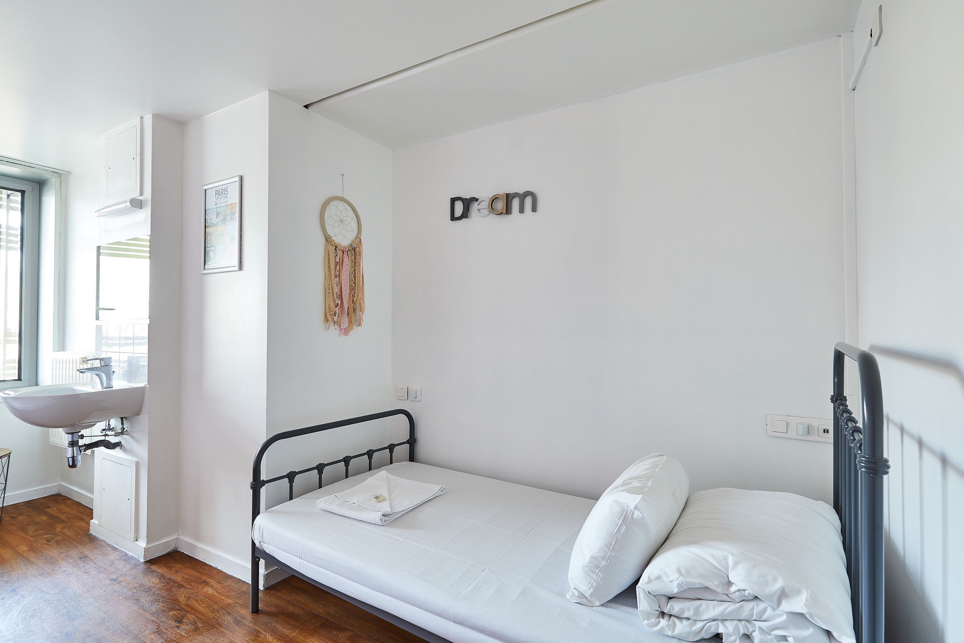 A small, clean room with a single bed, a white wall with a dream sign, a white sink, and a window.