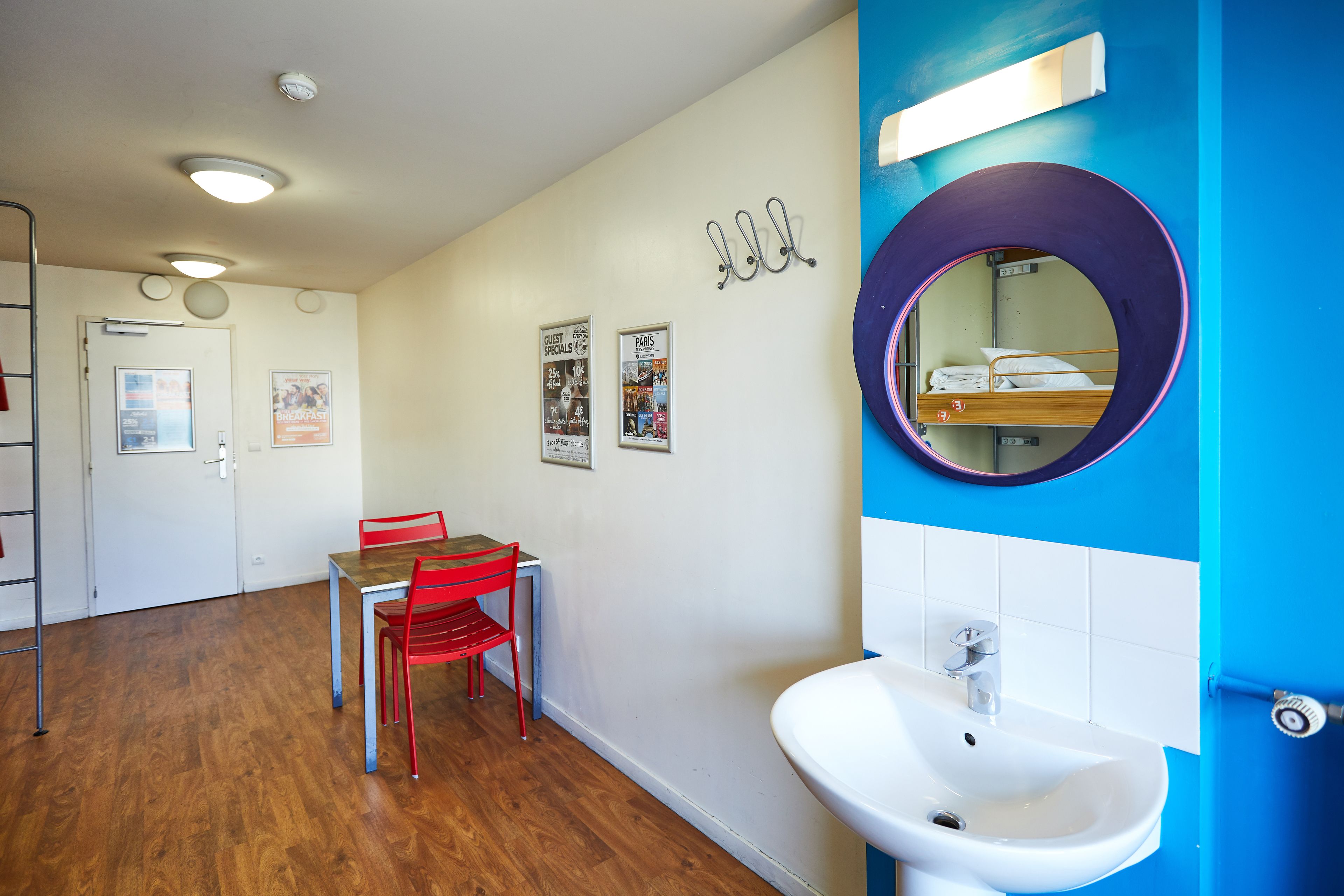A hotel room with a wooden floor, a dining table with red chairs, a white sink, and a mirror with a purple frame.