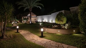 A nighttime view of a landscaped garden with a pathway, palm trees, and a building with illuminated exterior lighting. - Daphnis Villas (Platanias)