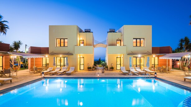 A swimming pool with lounge chairs and umbrellas, surrounded by buildings with balconies and windows. - Daphnis Villas (Platanias)